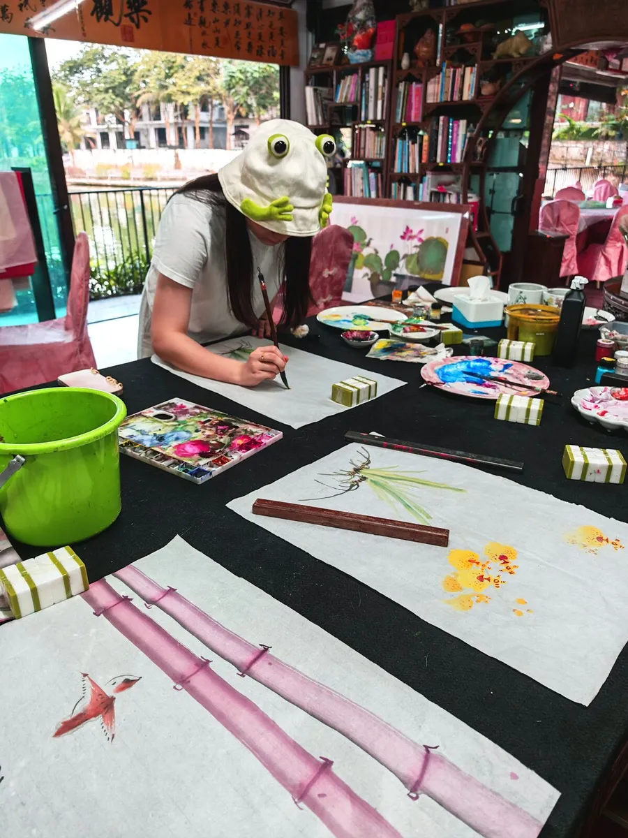 Hands painting bamboo in ink on rice paper at the big table, the lake beyond the window