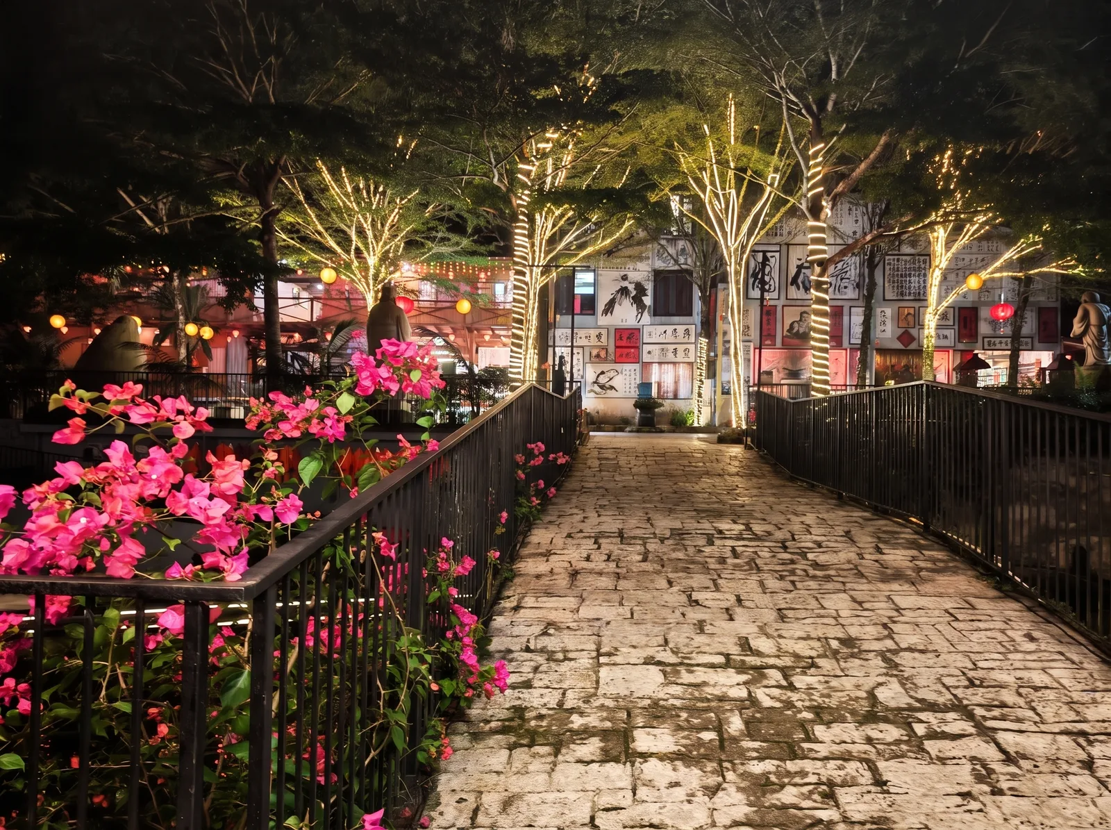 The lakeside walk at night: the trees wrapped in warm white-gold fairy lights, pink bougainvillea along the railing, the wall of framed paintings beyond